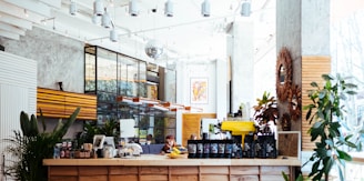 A modern, well-lit coffee shop interior with a wooden counter displaying various coffee bags and a yellow espresso machine. The space features plants, stylish light fixtures, and a mix of industrial and rustic decor. Large windows allow natural light to flood in, creating a bright and inviting atmosphere.