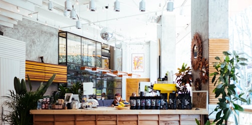 A modern, well-lit coffee shop interior with a wooden counter displaying various coffee bags and a yellow espresso machine. The space features plants, stylish light fixtures, and a mix of industrial and rustic decor. Large windows allow natural light to flood in, creating a bright and inviting atmosphere.