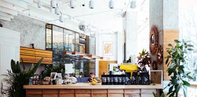 A modern, well-lit coffee shop interior with a wooden counter displaying various coffee bags and a yellow espresso machine. The space features plants, stylish light fixtures, and a mix of industrial and rustic decor. Large windows allow natural light to flood in, creating a bright and inviting atmosphere.
