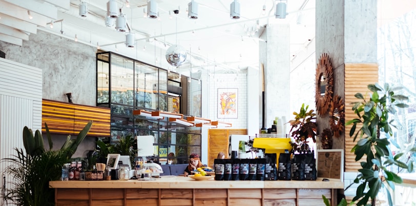 A modern, well-lit coffee shop interior with a wooden counter displaying various coffee bags and a yellow espresso machine. The space features plants, stylish light fixtures, and a mix of industrial and rustic decor. Large windows allow natural light to flood in, creating a bright and inviting atmosphere.