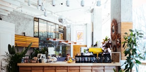 A modern, well-lit coffee shop interior with a wooden counter displaying various coffee bags and a yellow espresso machine. The space features plants, stylish light fixtures, and a mix of industrial and rustic decor. Large windows allow natural light to flood in, creating a bright and inviting atmosphere.