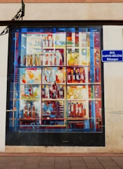 A mural painted on a wall depicting a colorful array of bottles and containers arranged on shelves. The painting uses an abstract style with vibrant splashes of color. A street sign reading 'Rue Sainte-Hélène Helengass' is attached to the wall next to the mural.