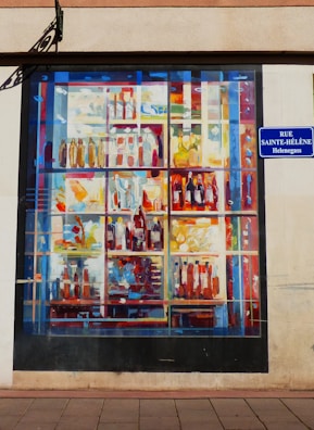 A mural painted on a wall depicting a colorful array of bottles and containers arranged on shelves. The painting uses an abstract style with vibrant splashes of color. A street sign reading 'Rue Sainte-Hélène Helengass' is attached to the wall next to the mural.