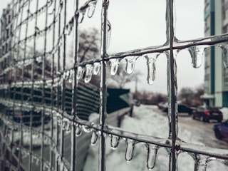 Icicles hang from a wire fence, with a blurred winter landscape in the background. Snow covers the ground, and a few parked cars and a building are visible, partially obscured by the icy pattern on the fence.