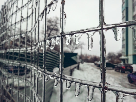 Icicles hang from a wire fence, with a blurred winter landscape in the background. Snow covers the ground, and a few parked cars and a building are visible, partially obscured by the icy pattern on the fence.