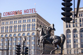A large, historic building with a prominent sign on the roof reading 'Congress Hotel' stands in the background. In the foreground, there is a bronze statue depicting a man wearing a headdress, riding a rearing horse. Traffic lights and light poles are also visible in the scene.