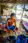 A man sits cross-legged on a bamboo platform beneath a thatched roof. He holds a smartphone and is surrounded by colorful items, including packets displayed behind him. The setting appears to be a tropical beach with visible sand, and there are makeshift structures in the background.
