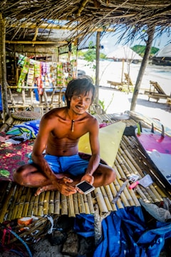 A man sits cross-legged on a bamboo platform beneath a thatched roof. He holds a smartphone and is surrounded by colorful items, including packets displayed behind him. The setting appears to be a tropical beach with visible sand, and there are makeshift structures in the background.