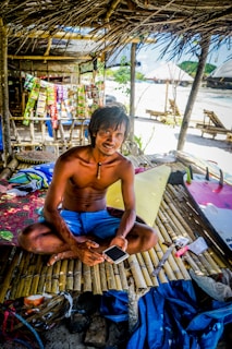 A man sits cross-legged on a bamboo platform beneath a thatched roof. He holds a smartphone and is surrounded by colorful items, including packets displayed behind him. The setting appears to be a tropical beach with visible sand, and there are makeshift structures in the background.