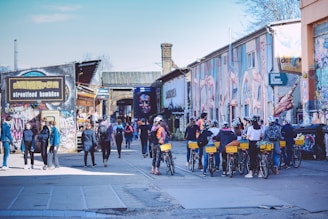 A lively urban scene with people walking and biking among buildings covered in colorful graffiti. A group of cyclists, some wearing helmets, are seen on the right. Pedestrians casually stroll on the left near a street food establishment. The atmosphere is vibrant with creative street art covering the walls.