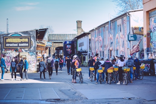 A lively urban scene with people walking and biking among buildings covered in colorful graffiti. A group of cyclists, some wearing helmets, are seen on the right. Pedestrians casually stroll on the left near a street food establishment. The atmosphere is vibrant with creative street art covering the walls.