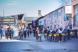 A lively urban scene with people walking and biking among buildings covered in colorful graffiti. A group of cyclists, some wearing helmets, are seen on the right. Pedestrians casually stroll on the left near a street food establishment. The atmosphere is vibrant with creative street art covering the walls.