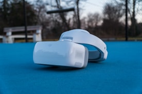 A pair of virtual reality goggles rests on a bright blue surface in an outdoor setting. In the background, there is a bench and a blurred view of trees under a cloudy sky.