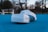 A pair of virtual reality goggles rests on a bright blue surface in an outdoor setting. In the background, there is a bench and a blurred view of trees under a cloudy sky.