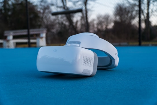 A pair of virtual reality goggles rests on a bright blue surface in an outdoor setting. In the background, there is a bench and a blurred view of trees under a cloudy sky.