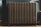 A vintage cast iron radiator stands against a paneled wall, showing intricate details and a tarnished patina. The radiator has multiple vertical sections and is positioned on a wooden floor with shadows cast from natural lighting.