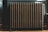 A vintage cast iron radiator stands against a paneled wall, showing intricate details and a tarnished patina. The radiator has multiple vertical sections and is positioned on a wooden floor with shadows cast from natural lighting.