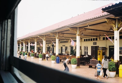An outdoor railway station platform with a colonial-style architecture featuring a long sheltered area supported by columns. Several people are moving through the platform, which is adorned with national flags. Potted plants are placed alongside the platform, and in the background, there are signs, benches, and an ATM.
