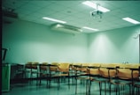 A classroom setting with several rows of empty orange and black chairs arranged in front of a wall-mounted projector. The room has a tiled ceiling with fluorescent lights and a wall air conditioning unit. A clock is visible on the wall, and the atmosphere appears clean and organized.
