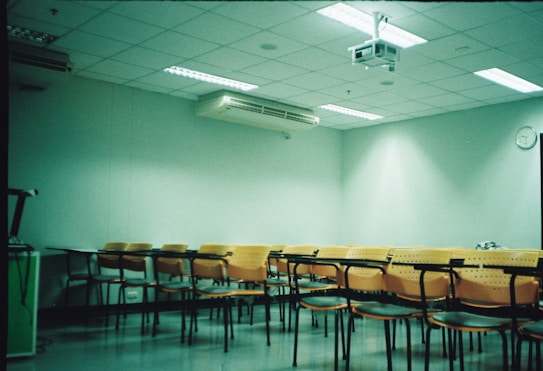 A classroom setting with several rows of empty orange and black chairs arranged in front of a wall-mounted projector. The room has a tiled ceiling with fluorescent lights and a wall air conditioning unit. A clock is visible on the wall, and the atmosphere appears clean and organized.