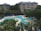 Aerial view of a resort-style pool area surrounded by lush green trees and plants. The pool features curves and multiple sections, with blue lounge chairs neatly arranged along the edges. In the background, a multi-story residential building with balconies overlooks the pool area. Blue umbrellas provide shade, and a few people can be seen enjoying the leisure space.