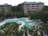 Aerial view of a resort-style pool area surrounded by lush green trees and plants. The pool features curves and multiple sections, with blue lounge chairs neatly arranged along the edges. In the background, a multi-story residential building with balconies overlooks the pool area. Blue umbrellas provide shade, and a few people can be seen enjoying the leisure space.