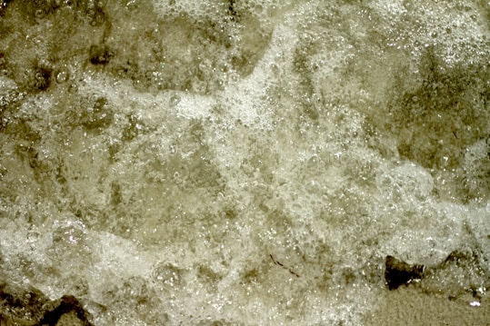 Bubbly water surfaces with foam covering most of the image, creating a textured appearance. The colors are mostly neutral, with hints of browns and whites.