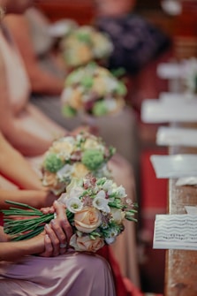 A group of people seated in a row, each holding a bouquet of flowers composed of roses, lilies, and greenery. The focus is on the flowers, with some individuals wearing pastel-colored dresses. The background is softly blurred, suggesting a formal setting, possibly a ceremony or event.