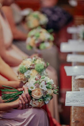 A group of people seated in a row, each holding a bouquet of flowers composed of roses, lilies, and greenery. The focus is on the flowers, with some individuals wearing pastel-colored dresses. The background is softly blurred, suggesting a formal setting, possibly a ceremony or event.
