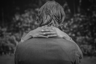 A person with long hair is seen from behind, embraced by another person whose hands with visible rings and bracelets are wrapped around the shoulders. The scene is captured in black and white, giving it a timeless and intimate feel.
