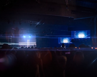 A dimly lit concert or event space with a stage illuminated by bright blue and white lights. The background features large screens displaying similar bright lights, creating a symmetrical pattern. A crowd of silhouetted people fills the foreground, suggesting a live music or entertainment event.