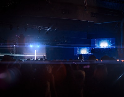 A dimly lit concert or event space with a stage illuminated by bright blue and white lights. The background features large screens displaying similar bright lights, creating a symmetrical pattern. A crowd of silhouetted people fills the foreground, suggesting a live music or entertainment event.