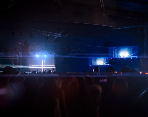 A dimly lit concert or event space with a stage illuminated by bright blue and white lights. The background features large screens displaying similar bright lights, creating a symmetrical pattern. A crowd of silhouetted people fills the foreground, suggesting a live music or entertainment event.