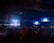 A dimly lit concert or event space with a stage illuminated by bright blue and white lights. The background features large screens displaying similar bright lights, creating a symmetrical pattern. A crowd of silhouetted people fills the foreground, suggesting a live music or entertainment event.
