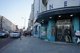 A street view featuring a cinema with large letters spelling 'CINEMA' on its facade. The building has a round, blue-colored entrance. There are several people walking along the sidewalk, and one person standing near the entrance. Various cars are lined up along the street, with buildings stretching into the distance. The scene takes place during the day with a clear sky overhead.