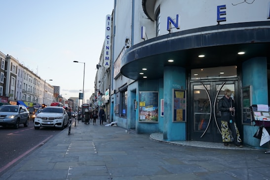 A street view featuring a cinema with large letters spelling 'CINEMA' on its facade. The building has a round, blue-colored entrance. There are several people walking along the sidewalk, and one person standing near the entrance. Various cars are lined up along the street, with buildings stretching into the distance. The scene takes place during the day with a clear sky overhead.