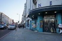 A street view featuring a cinema with large letters spelling 'CINEMA' on its facade. The building has a round, blue-colored entrance. There are several people walking along the sidewalk, and one person standing near the entrance. Various cars are lined up along the street, with buildings stretching into the distance. The scene takes place during the day with a clear sky overhead.