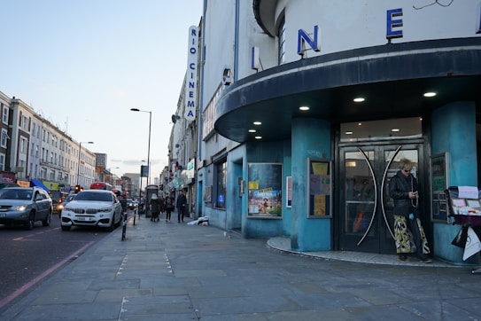 A street view featuring a cinema with large letters spelling 'CINEMA' on its facade. The building has a round, blue-colored entrance. There are several people walking along the sidewalk, and one person standing near the entrance. Various cars are lined up along the street, with buildings stretching into the distance. The scene takes place during the day with a clear sky overhead.