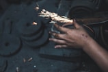 A close-up of a person's hand holding a tool against a grinding wheel, producing bright orange sparks. The hand appears to have some scars or marks. The background is dark and out of focus.