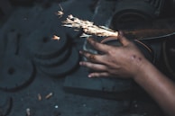 A close-up of a person's hand holding a tool against a grinding wheel, producing bright orange sparks. The hand appears to have some scars or marks. The background is dark and out of focus.