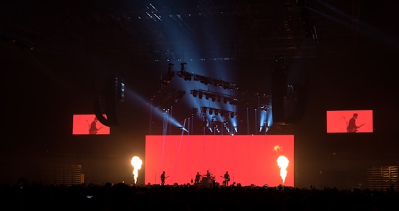 A dramatic stage performance with bright red lighting and fire columns, featuring a band with silhouetted musicians against a large red screen. Overhead lighting rig adds dynamic light effects, and additional screens display band members.