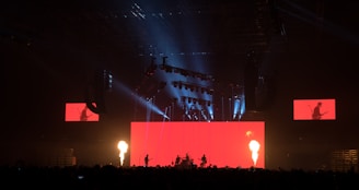 A dramatic stage performance with bright red lighting and fire columns, featuring a band with silhouetted musicians against a large red screen. Overhead lighting rig adds dynamic light effects, and additional screens display band members.