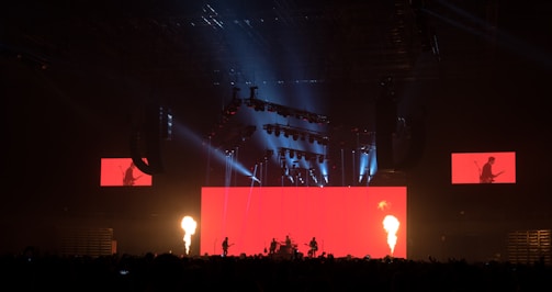 A dramatic stage performance with bright red lighting and fire columns, featuring a band with silhouetted musicians against a large red screen. Overhead lighting rig adds dynamic light effects, and additional screens display band members.