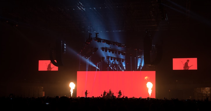 A dramatic stage performance with bright red lighting and fire columns, featuring a band with silhouetted musicians against a large red screen. Overhead lighting rig adds dynamic light effects, and additional screens display band members.