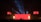 A dramatic stage performance with bright red lighting and fire columns, featuring a band with silhouetted musicians against a large red screen. Overhead lighting rig adds dynamic light effects, and additional screens display band members.