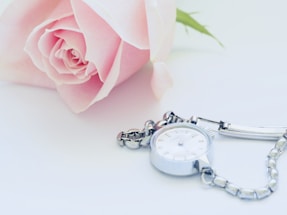 A close-up of a delicate women's watch resting beside a vintage letter and a soft pink rose.
