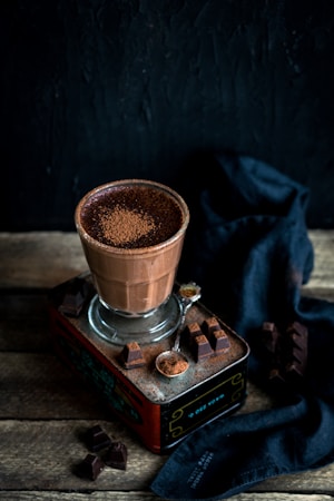 A glass filled with a rich, frothy chocolate drink sits on a vintage tin box. The surrounding area is decorated with pieces of dark chocolate and a small spoon filled with cocoa powder. A dark cloth adds texture and contrast in the background.