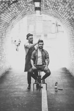 A hairstylist dressed in casual attire and a hat is performing a haircut on a seated man in an urban tunnel. The scene is captured in black and white, highlighting the arched brickwork of the tunnel. Hair products are visible on the ground beside the stylist.