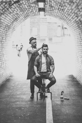 A hairstylist dressed in casual attire and a hat is performing a haircut on a seated man in an urban tunnel. The scene is captured in black and white, highlighting the arched brickwork of the tunnel. Hair products are visible on the ground beside the stylist.