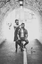 A hairstylist dressed in casual attire and a hat is performing a haircut on a seated man in an urban tunnel. The scene is captured in black and white, highlighting the arched brickwork of the tunnel. Hair products are visible on the ground beside the stylist.
